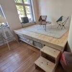 Schmigalla Wohnen Hobby Ein helles Zimmer mit einem großen, maßgefertigten Holzpodest, das als Wohnbereich mit Tisch und Stühlen dient und unter dem ein ausziehbares Bett verstaut ist.