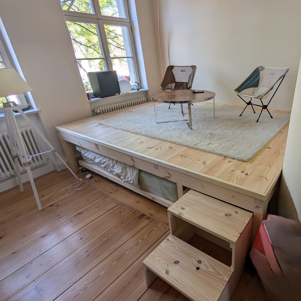 Schmigalla Wohnen Hobby Ein helles Zimmer mit einem großen, maßgefertigten Holzpodest, das als Wohnbereich mit Tisch und Stühlen dient und unter dem ein ausziehbares Bett verstaut ist.