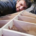 Galerie Schmigalla Wohnen Hobby. Nahaufnahme einer hölzernen Rippenkonstruktion aus Sperrholz, über die sich ein Mann konzentriert beugt, um die Ausrichtung der Spanten zu prüfen.