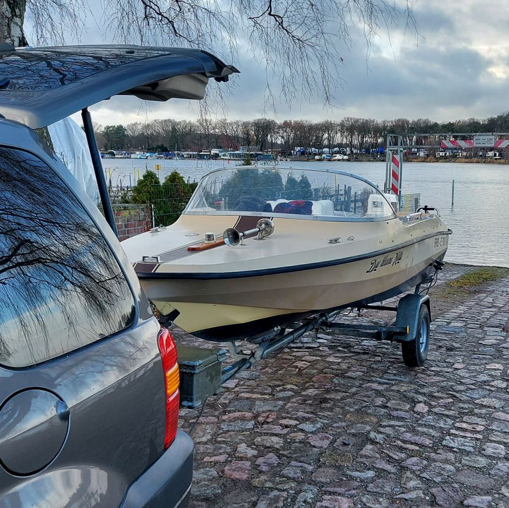 Hobby. Ein hellbeiges Oldtimer-Motorboot mit großer Windschutzscheibe und markanter Chromhupe auf einem Trailer. Das Boot steht auf einer Slipanlage aus Kopfsteinpflaster am Ufer eines Sees, angekoppelt an einen grauen Pkw.