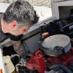 Ein Mann im schwarzen T-Shirt beugt sich über einen rot-schwarzen Bootsmotor in einem weißen Motorboot, um Reparaturen oder Wartungsarbeiten durchzuführen.