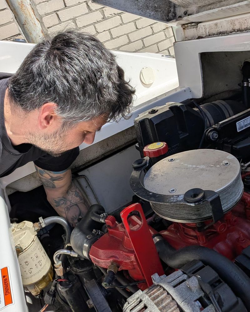 Ein Mann im schwarzen T-Shirt beugt sich über einen rot-schwarzen Bootsmotor in einem weißen Motorboot, um Reparaturen oder Wartungsarbeiten durchzuführen.