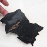 Eine Hand hält ein großes, flaches Stück abgeplatztes schwarzes Antifouling vor einem weißen Bootsrumpf.